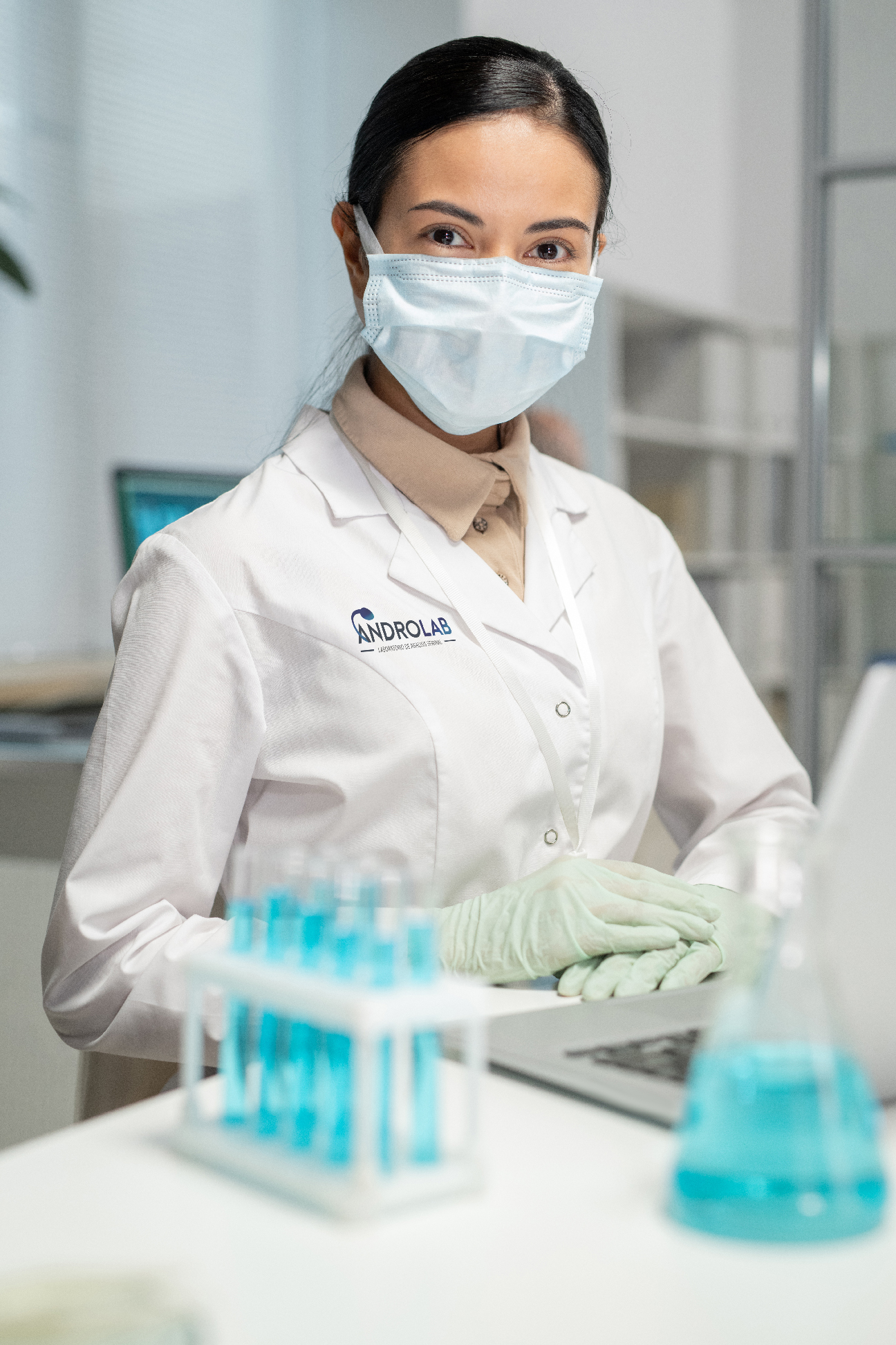 Young female scientist in whitecoat, gloves and protective mask sitting by desk in front of laptop and set of flasks with blue liquid